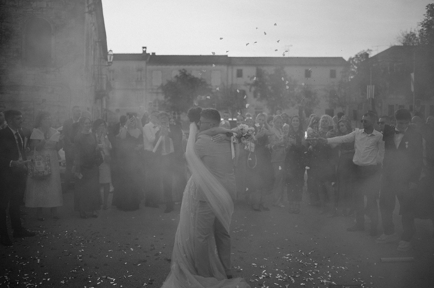 Black and white photo of a bride and groom kissing surrounded by wedding guests outdoors.
