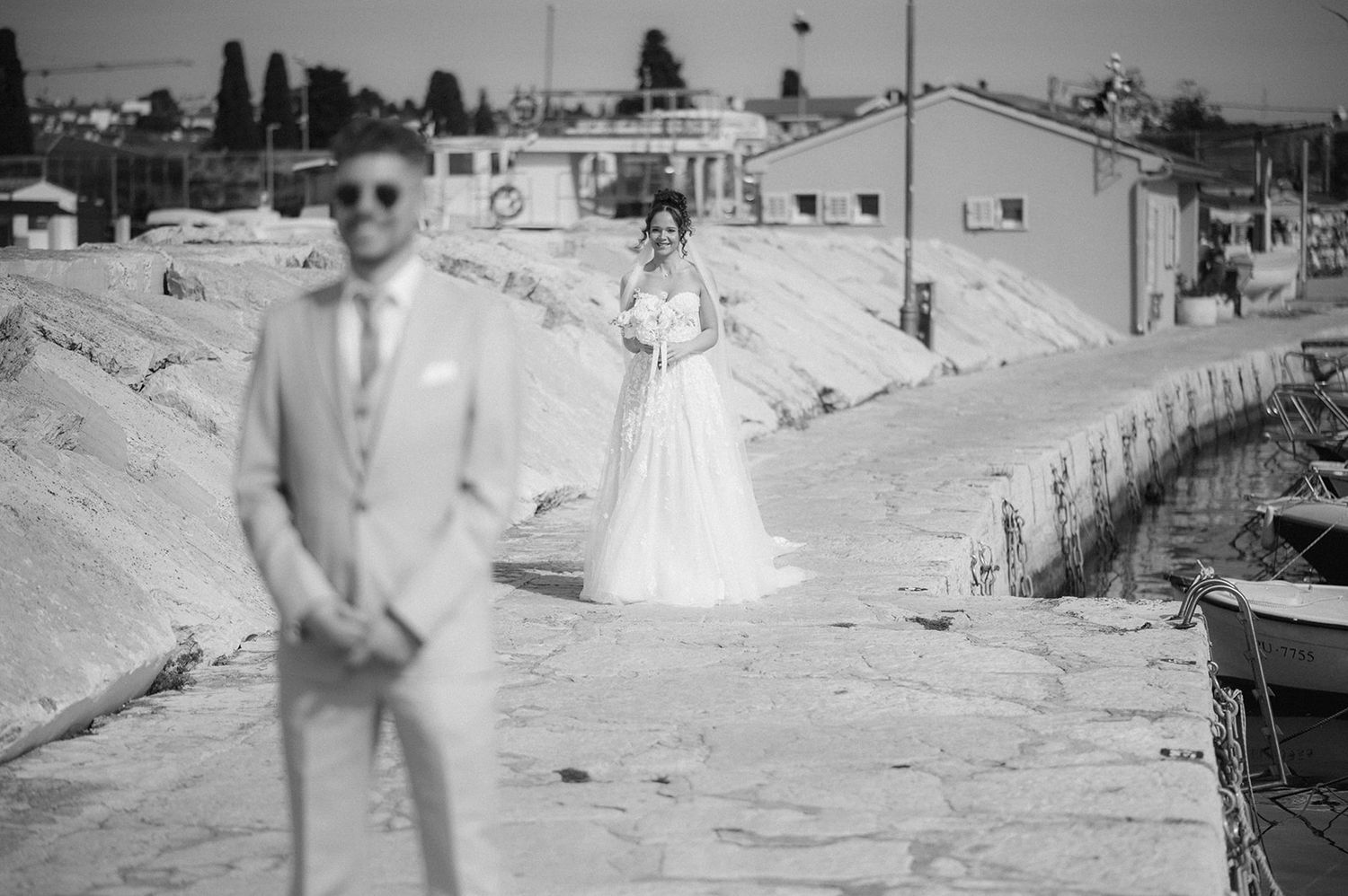 Black and white wedding photo of bride in white gown approaching groom in suit on a seaside pier.