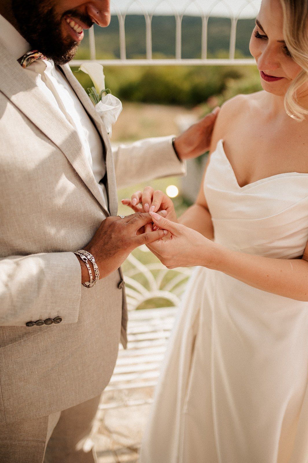 Bride placing wedding ring on groom's finger during outdoor ceremony, couple in elegant wedding attire.