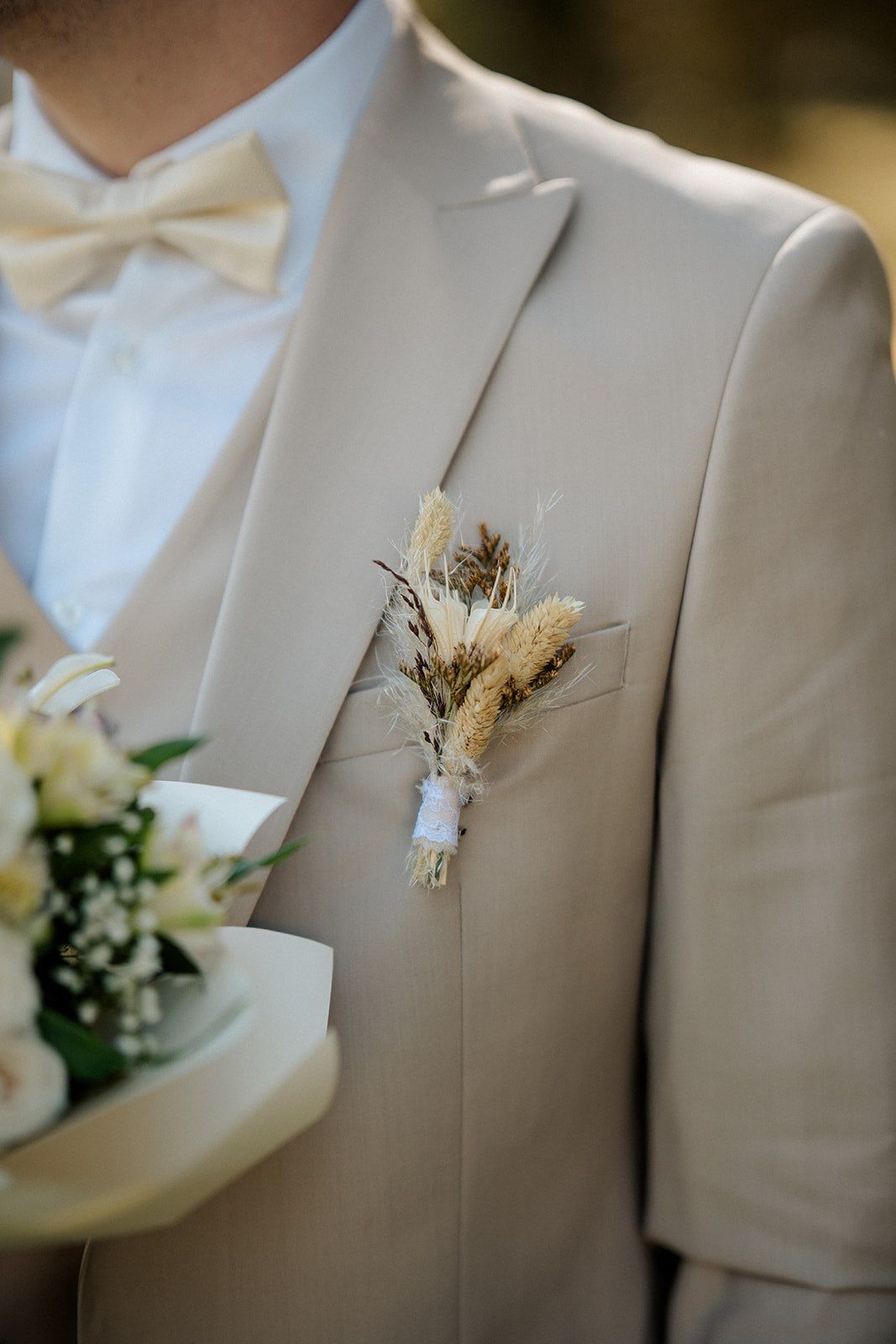 Groom in beige suit with dried floral boutonniere and bow tie holding a white wedding bouquet.