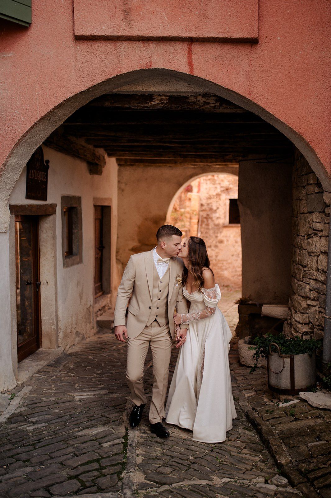 Bride and groom kissing under a stone arch in a charming European cobblestone alley, groom in beige suit.