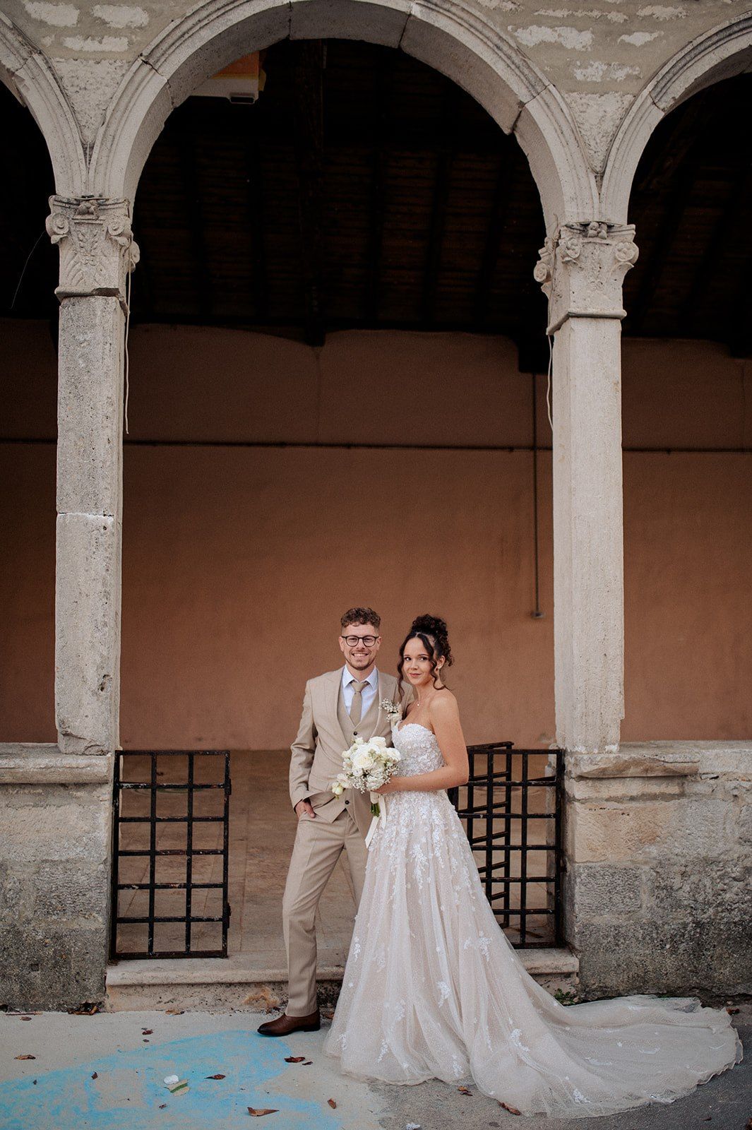 Bride and groom posing under stone arches at a historic venue, bride holding white bouquet in elegant lace gown.