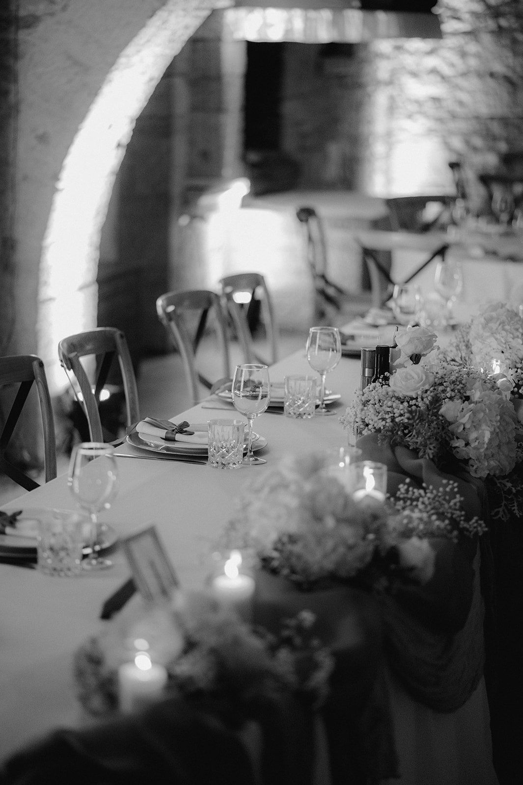 Black and white photo of an elegant wedding reception table with floral centerpieces, candles, and glassware in a rustic venue.
