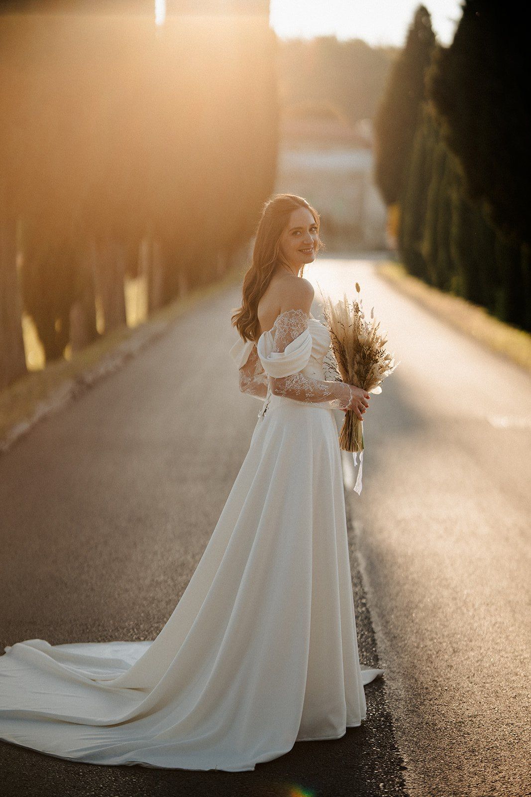 Bride in white off-shoulder gown with lace sleeves holds wheat bouquet on tree-lined road at golden sunset.
