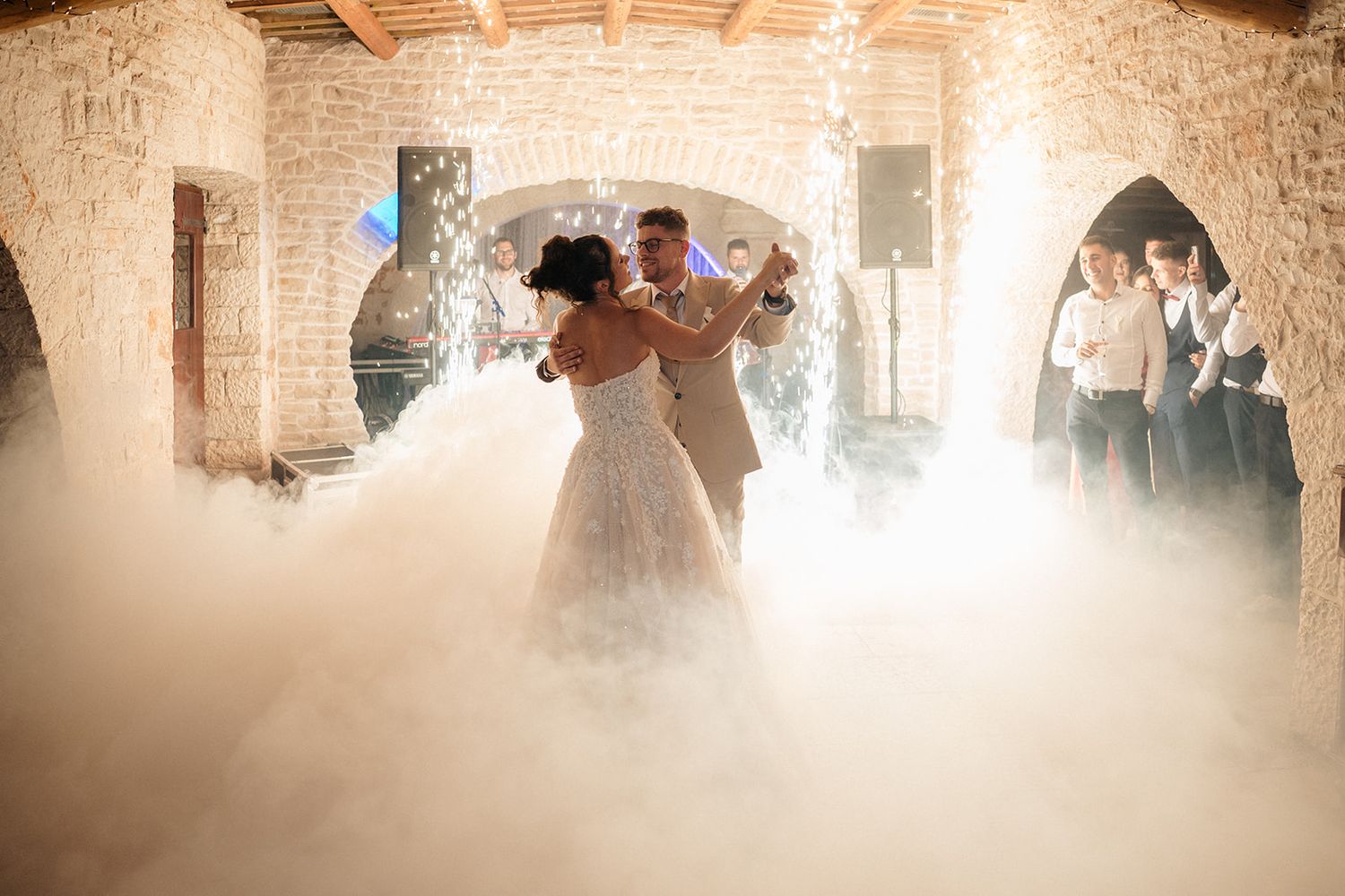 Bride and groom share first dance surrounded by dramatic fog machine clouds in a rustic stone wedding venue.