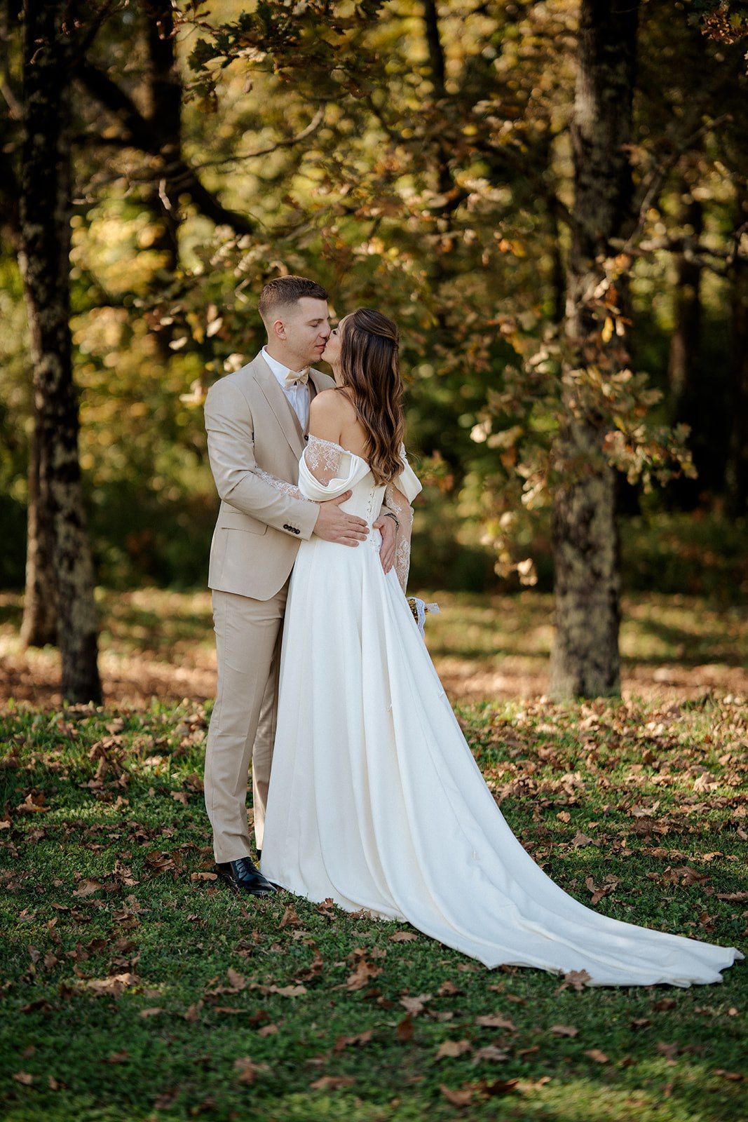 A bride and groom share a romantic kiss in a sunlit autumn forest, she in a flowing white gown, he in a tan suit.
