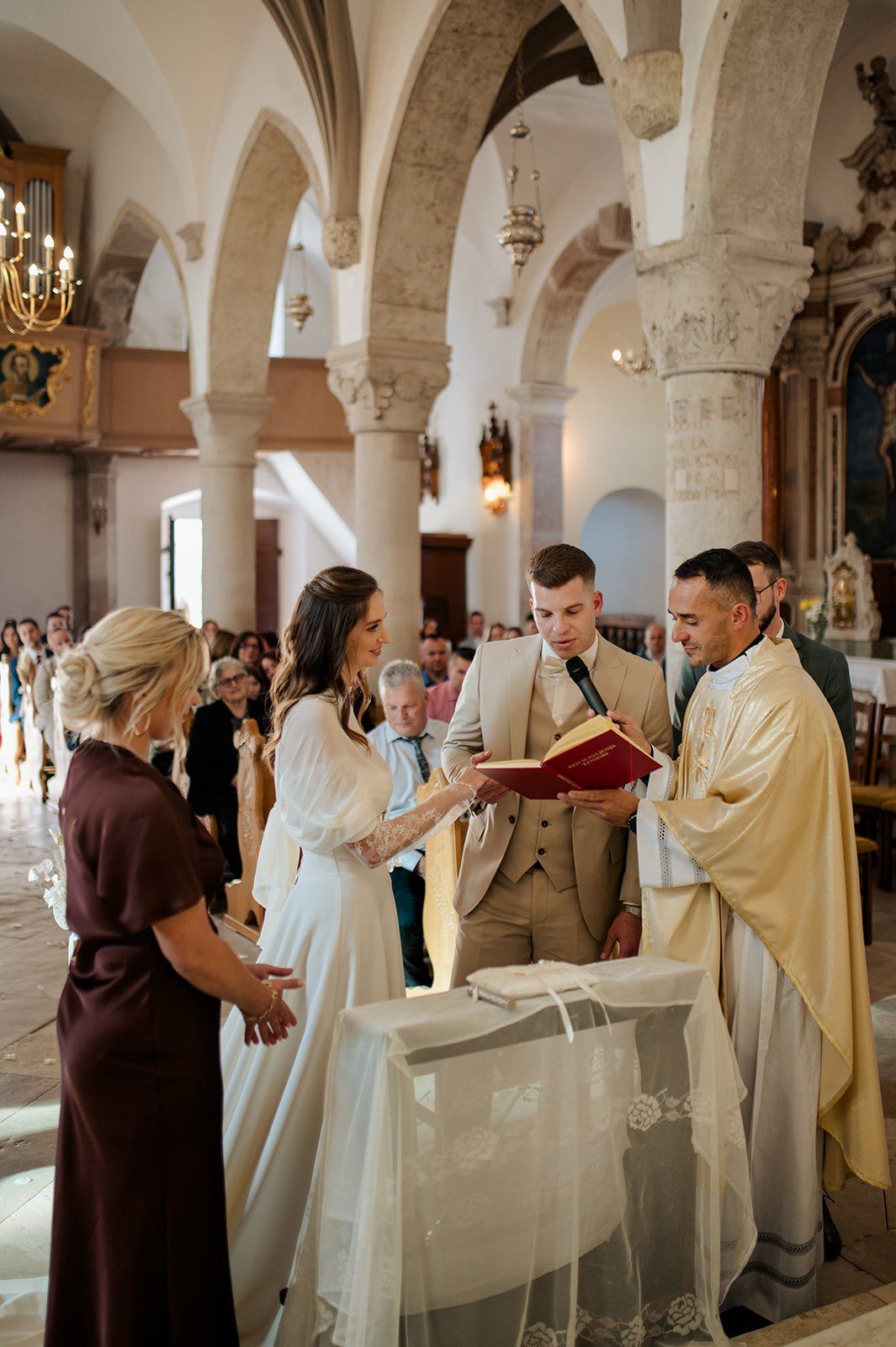 Bride and groom exchange vows with a priest in an elegant stone church during a traditional wedding ceremony.