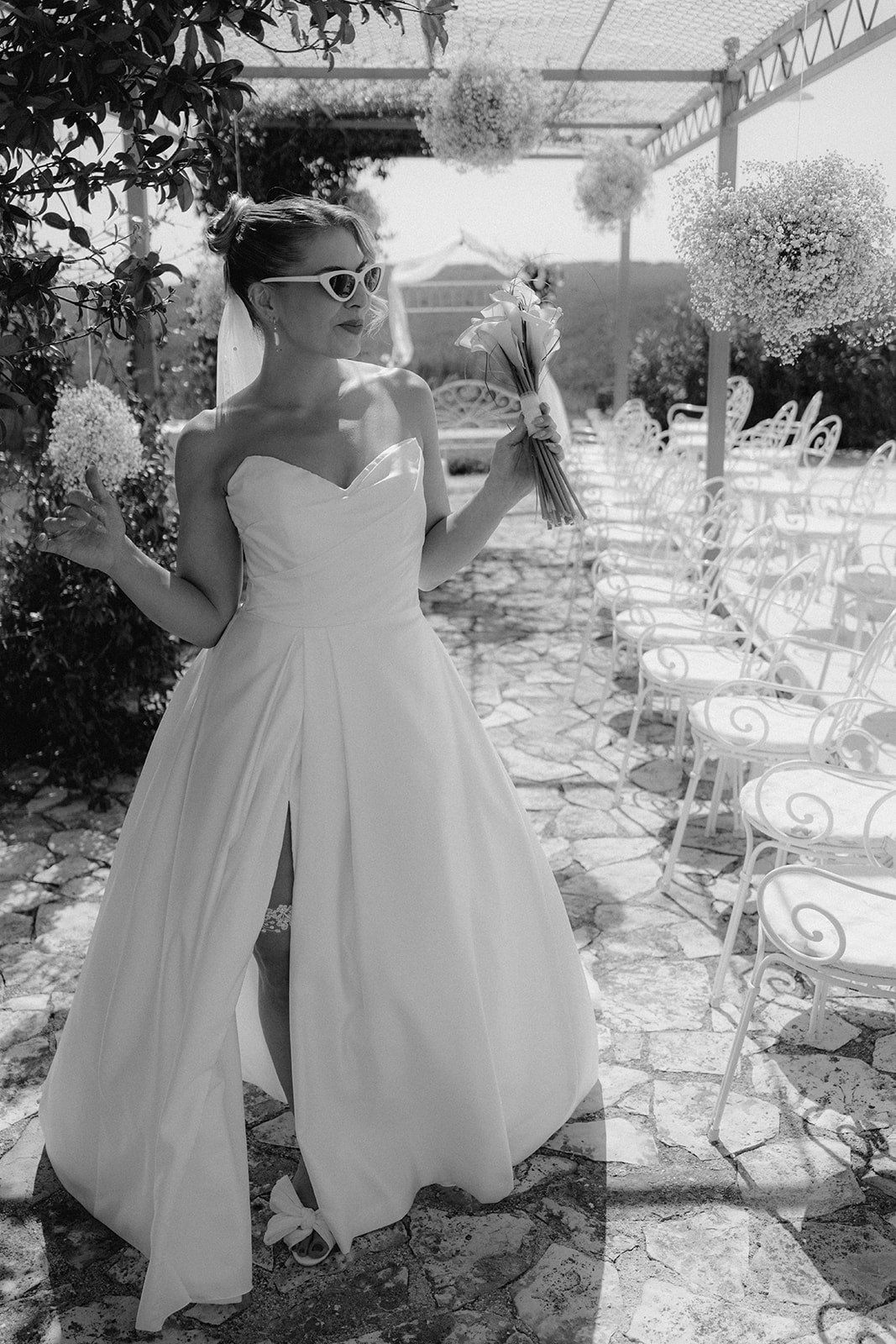 Bride in white strapless gown with sunglasses standing by outdoor reception table in black and white wedding photography.