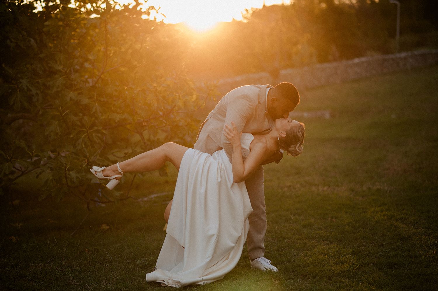 Couple sharing a romantic dip kiss at golden sunset in lush garden, bride in white gown, groom in tan suit.