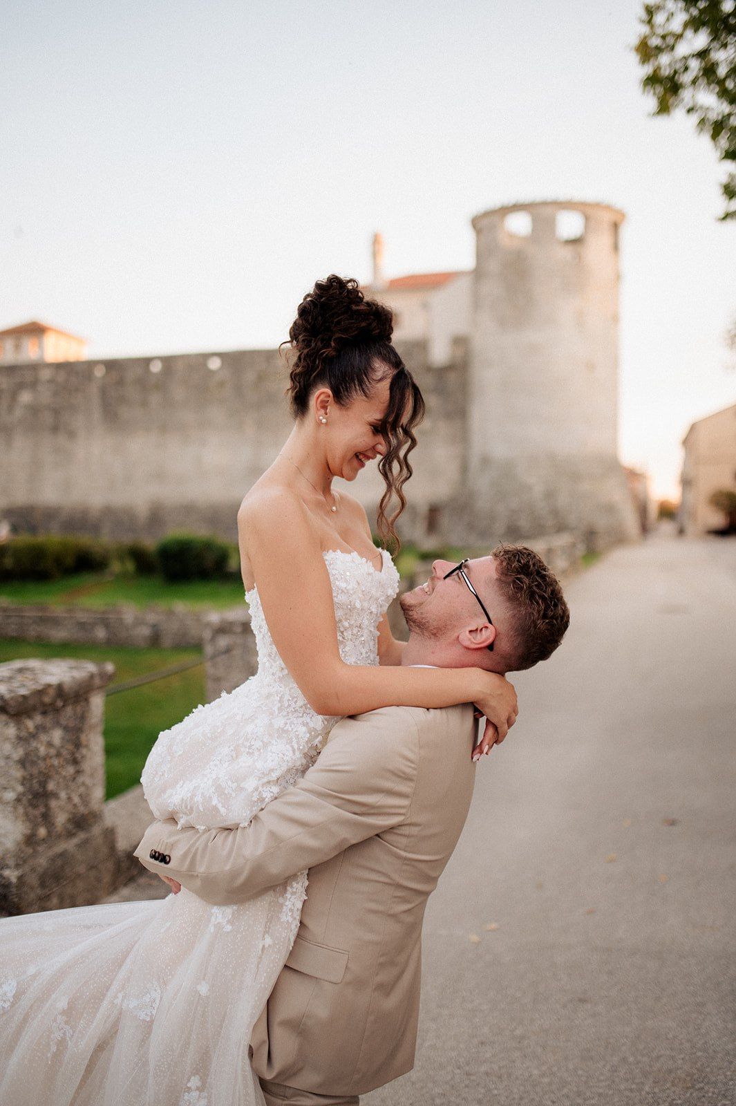 Groom lifts bride in white floral gown near ancient stone walls at sunset during romantic wedding celebration.