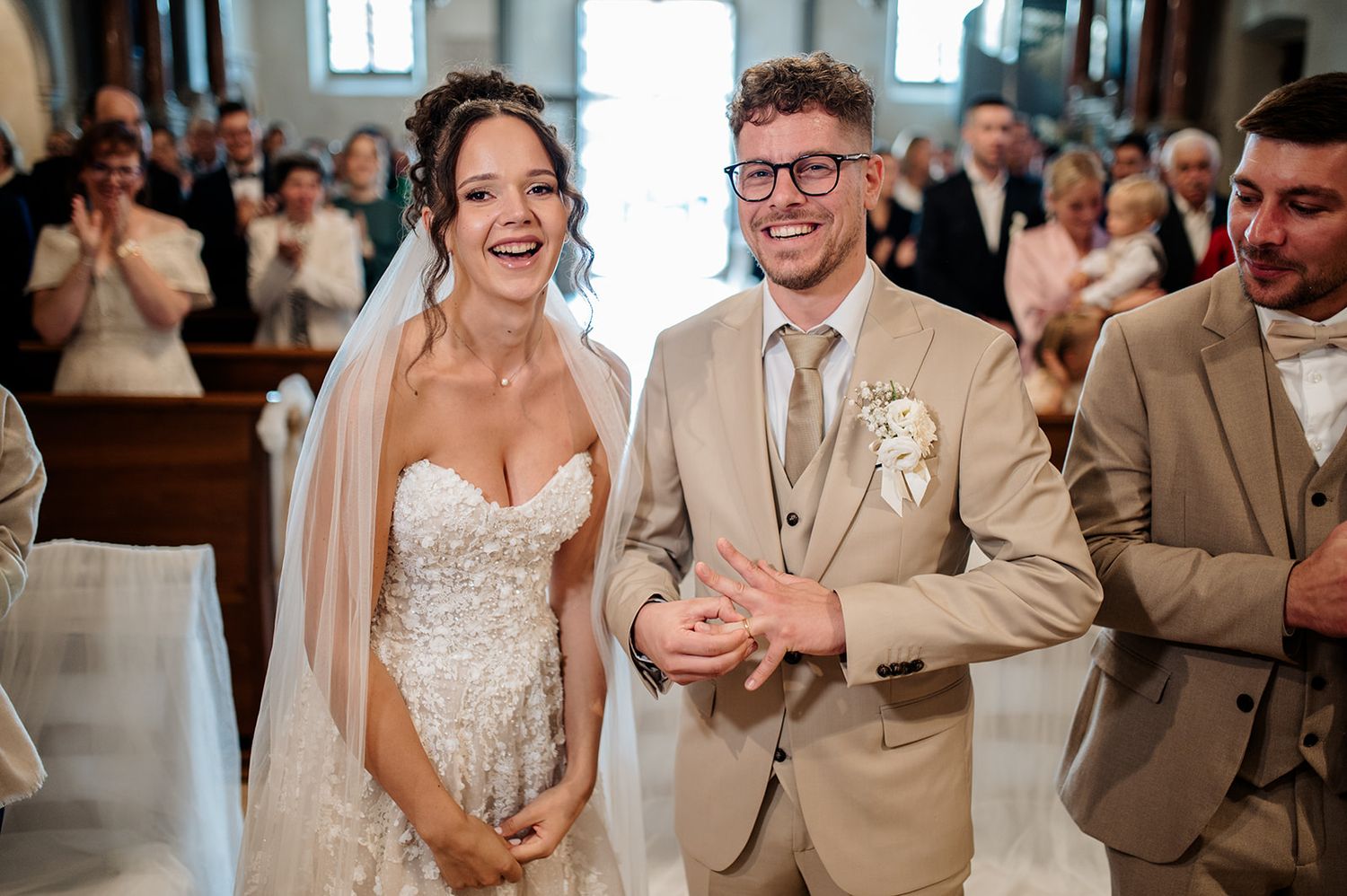 Happy bride and groom exchanging rings at church altar during wedding ceremony, guests applauding in background.