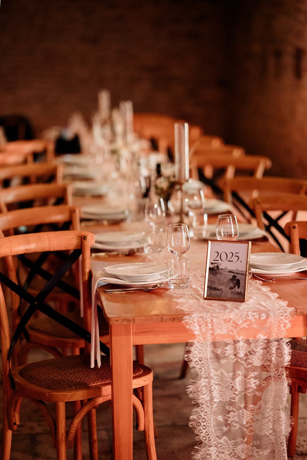 Elegant wedding reception table with lace runner, wine glasses, white plates, and a 2025 table number card in a rustic venue.