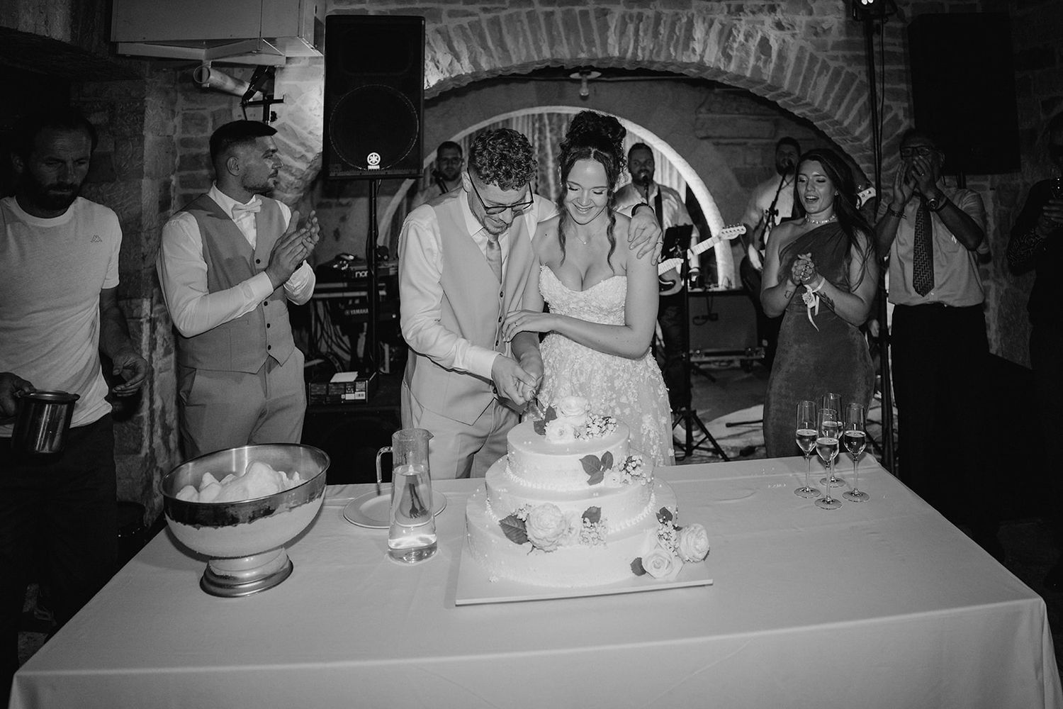 Black and white photo of a bride and groom cutting their wedding cake surrounded by guests at a rustic venue.