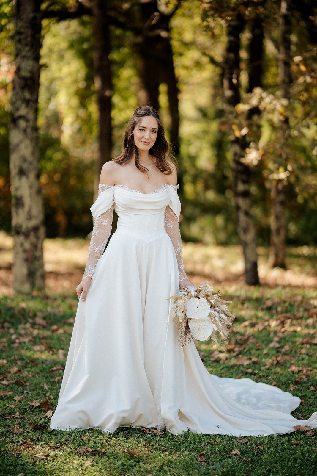 Bride in off-shoulder white A-line gown holding dried floral bouquet, posing in autumn forest setting.