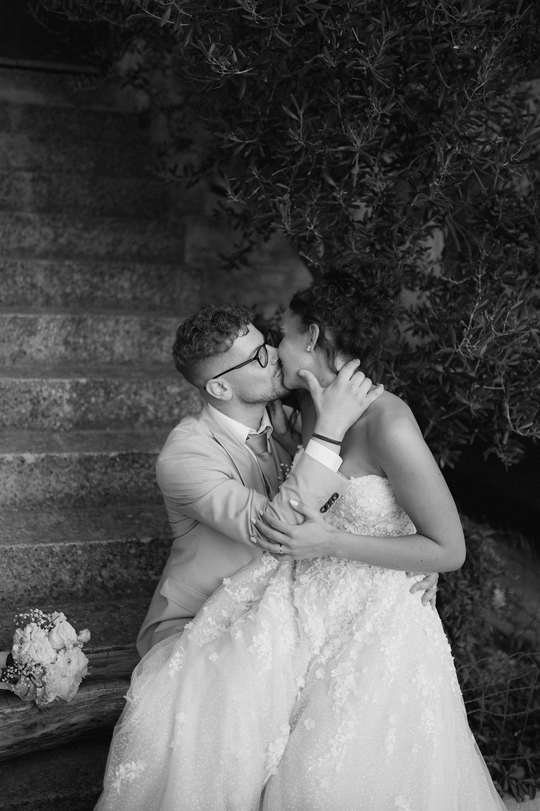 Black and white romantic photo of bride and groom kissing on stone steps with bouquet nearby.