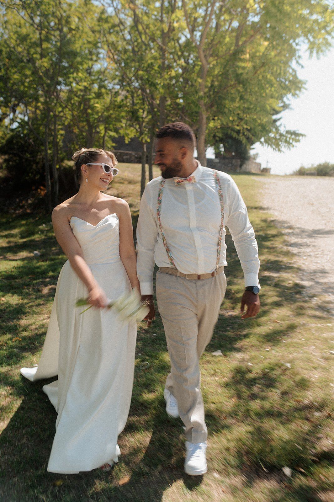 A bride and groom walk hand-in-hand outdoors on their wedding day, smiling at each other in elegant attire.