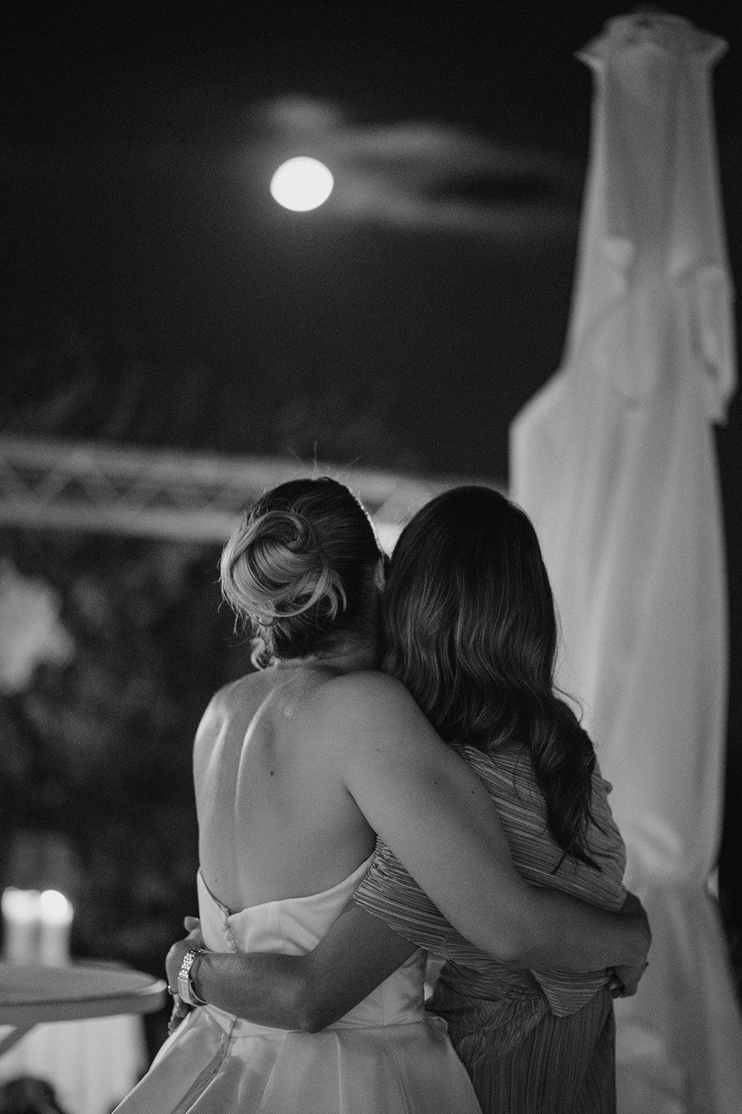 Black and white photo of bride and guest embracing under full moon at outdoor evening wedding reception.