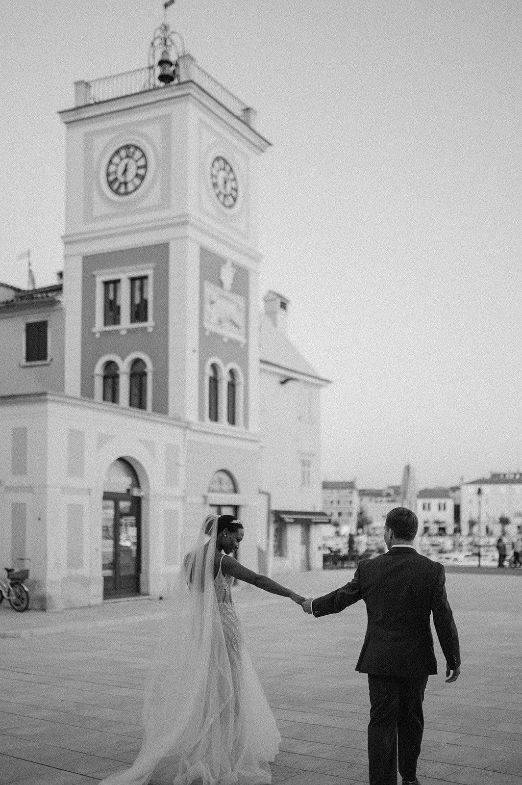 Newlyweds holding hands in front of Italian clock tower in black and white photography.