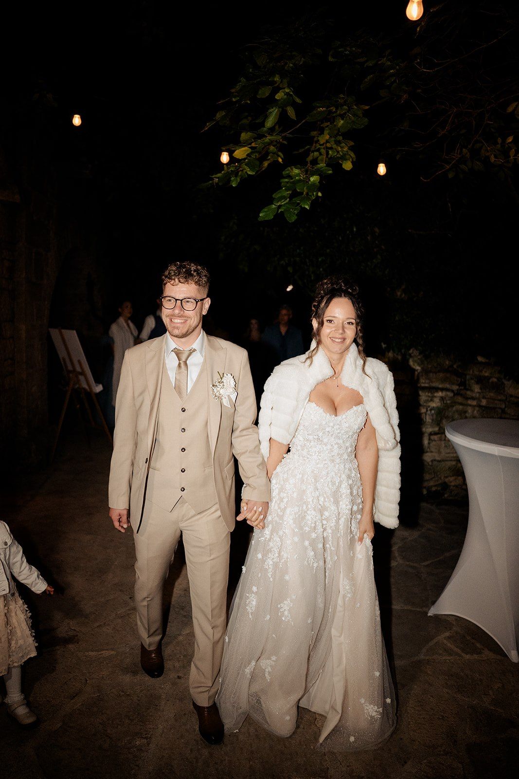 Bride in white lace gown and groom in tan suit hold hands smiling at outdoor evening wedding reception.