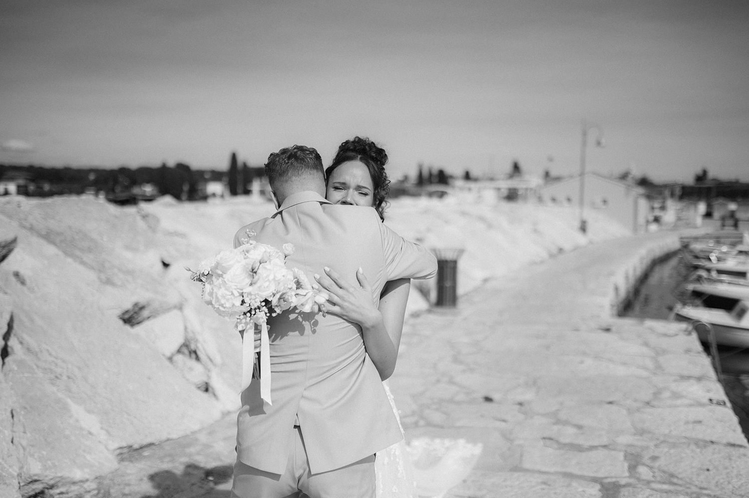 Bride and groom share a tender embrace on a rocky harbor pier, with the bride holding a white bouquet.