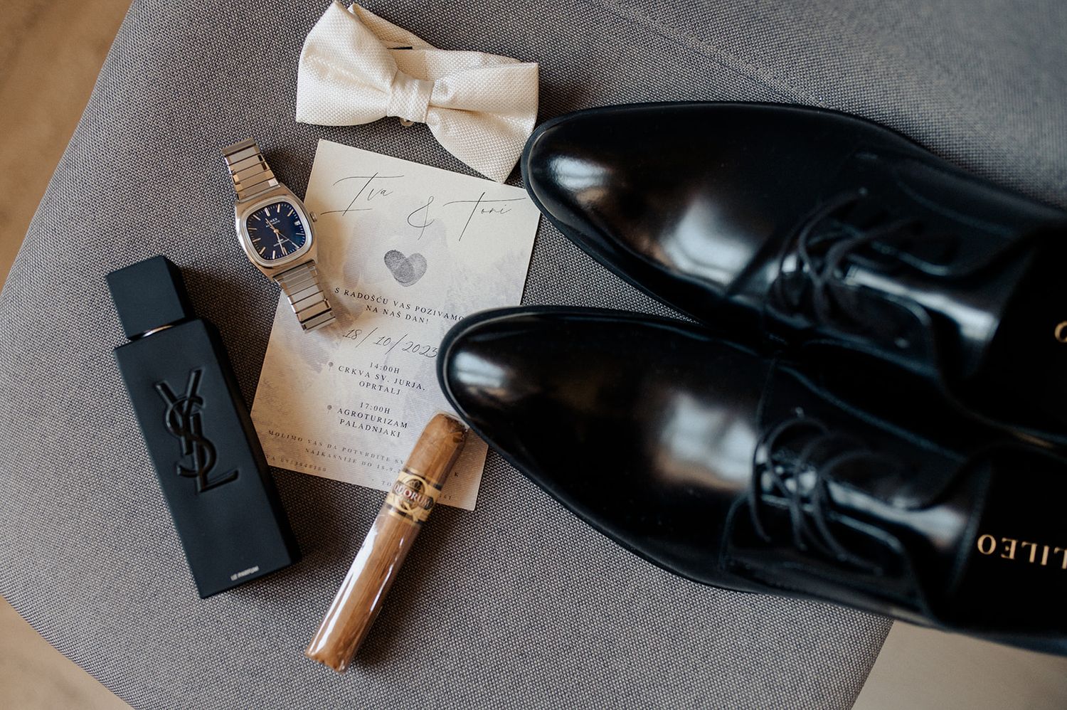 Black dress shoes, YSL cologne, gold watch, bow tie, and wedding invitation styled as groom flat lay accessories.