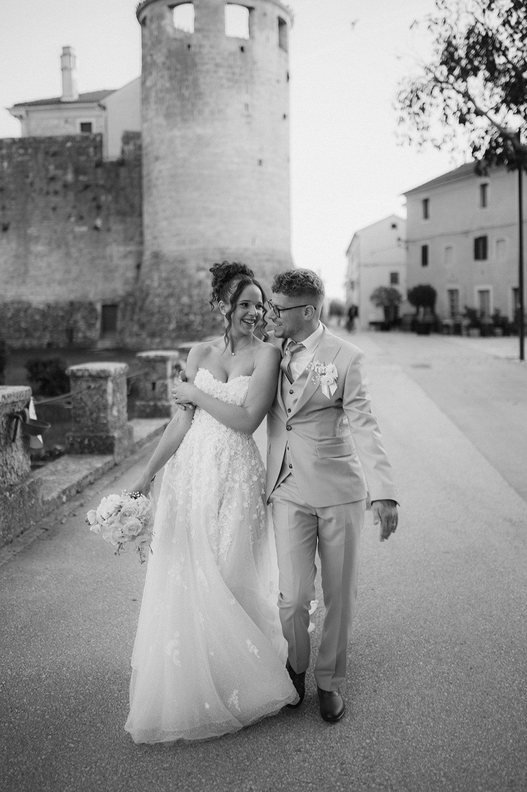 Bride and groom walking together in black and white wedding photo near historic stone tower.