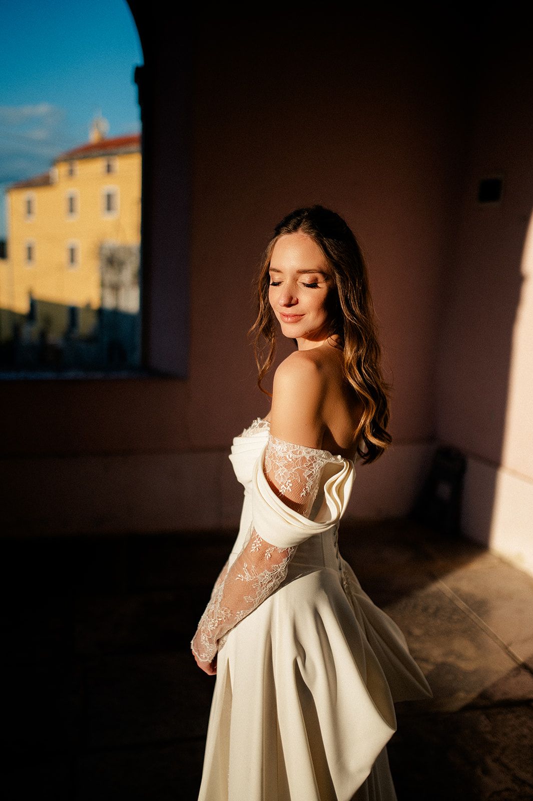 Bride in off-shoulder white lace gown poses gracefully in golden sunlight against a pink wall.