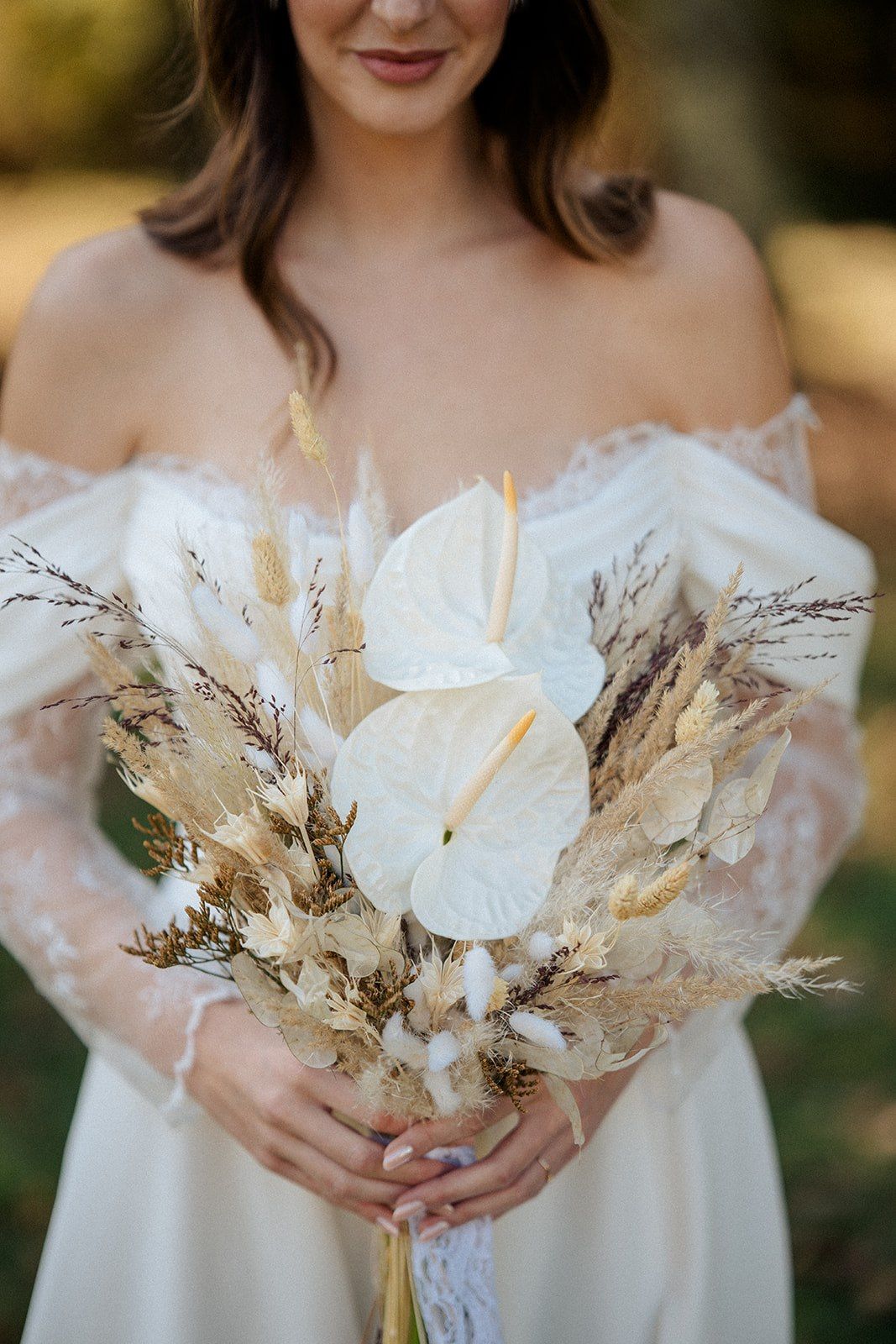 Bride in off-shoulder lace gown holding boho dried flower bouquet with white anthuriums and pampas grass.