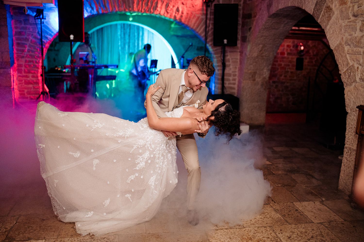 Bride and groom share romantic first dance dip in a stone venue with colorful lights and dry ice fog.