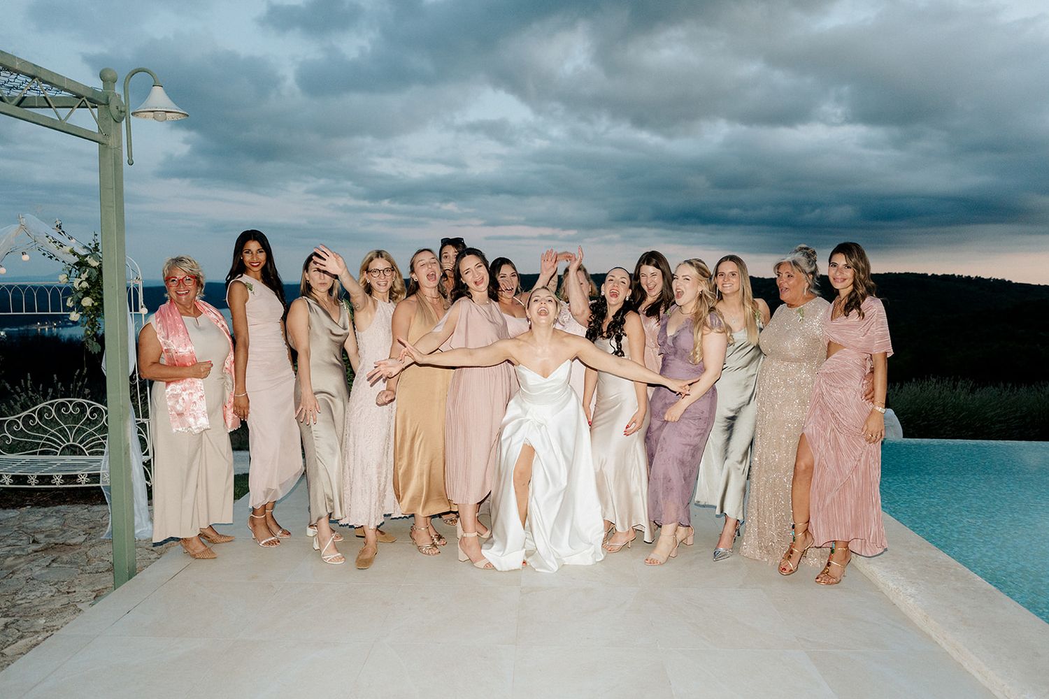 Bride poses with bridesmaids in pastel dresses by a pool under dramatic cloudy skies at outdoor wedding.