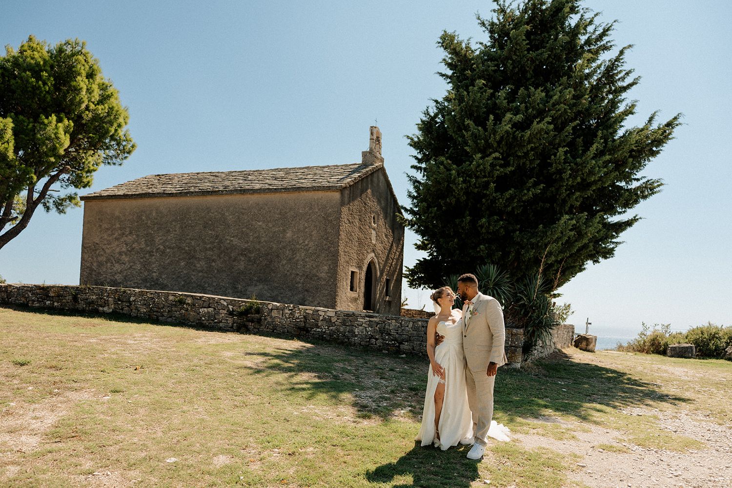 Bride and groom embrace outside a rustic stone chapel surrounded by lush green trees on a sunny day.