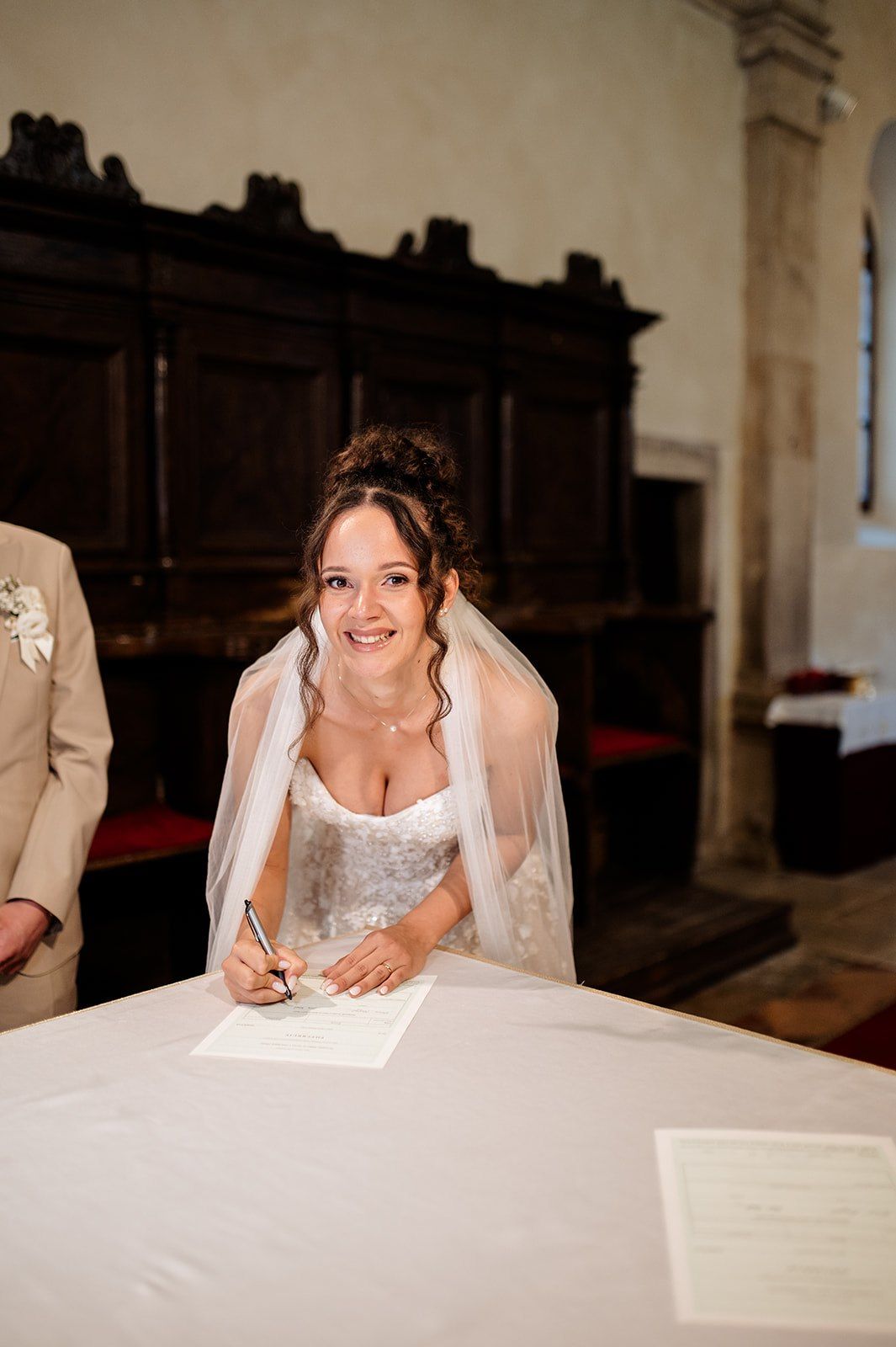 Happy bride in lace gown and veil signing wedding register at ceremony.
