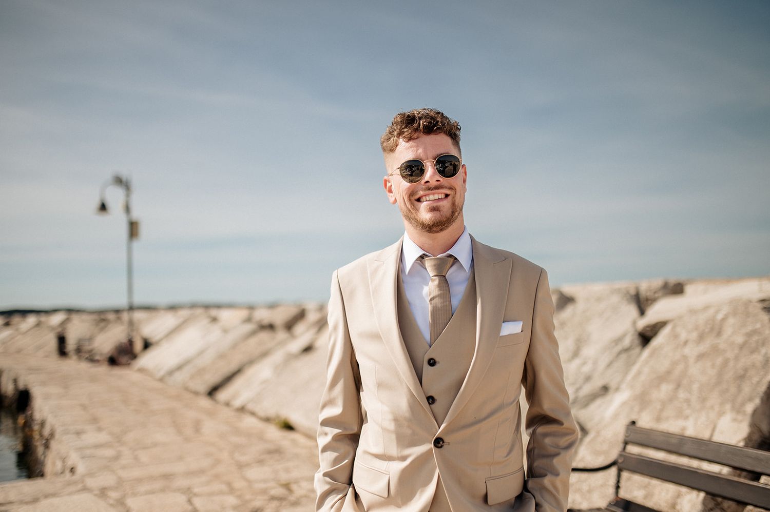 Smiling man in beige suit and sunglasses posing outdoors near a waterfront on a sunny day.