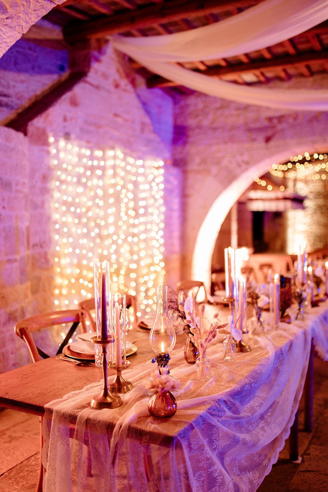 Elegant wedding reception table with candles, fairy lights, and lace runner in a rustic stone venue.