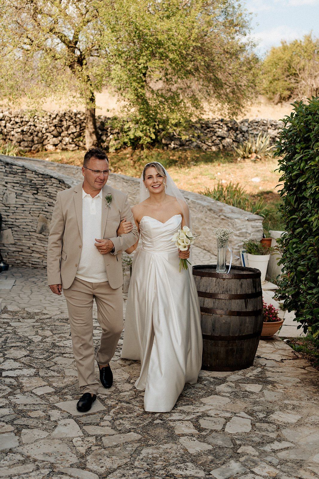 Bride in white gown walks arm-in-arm with man in tan suit on stone path at outdoor wedding venue.