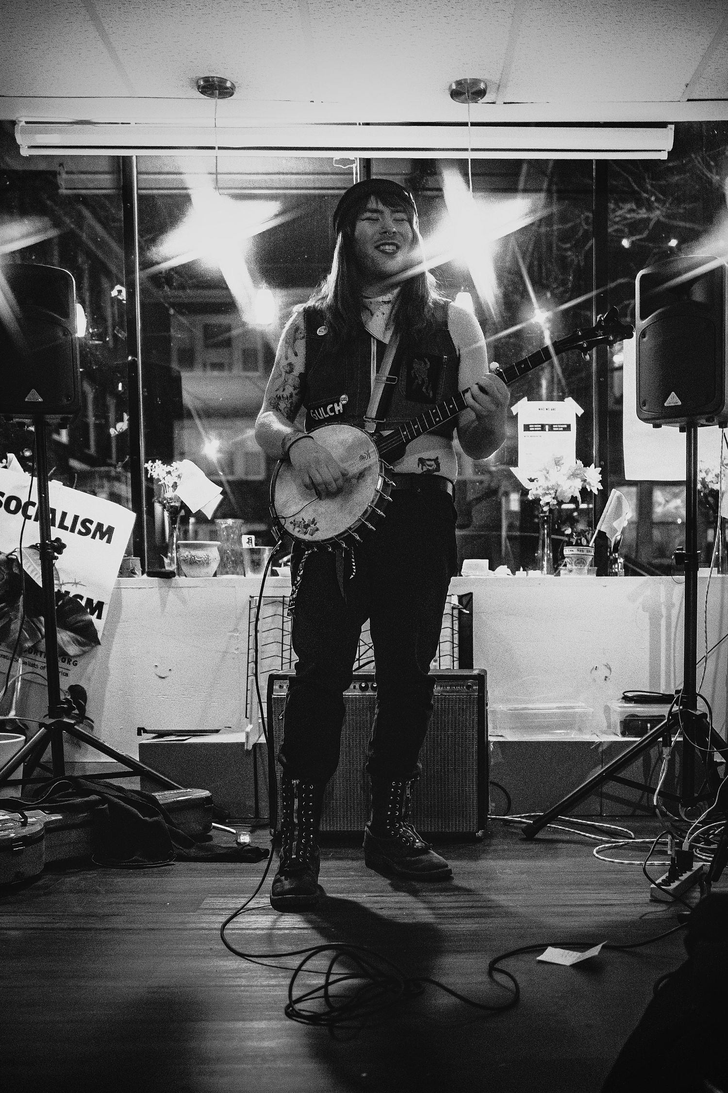 Black and white photo of a smiling musician playing banjo on stage with speakers and socialism sign in background.
