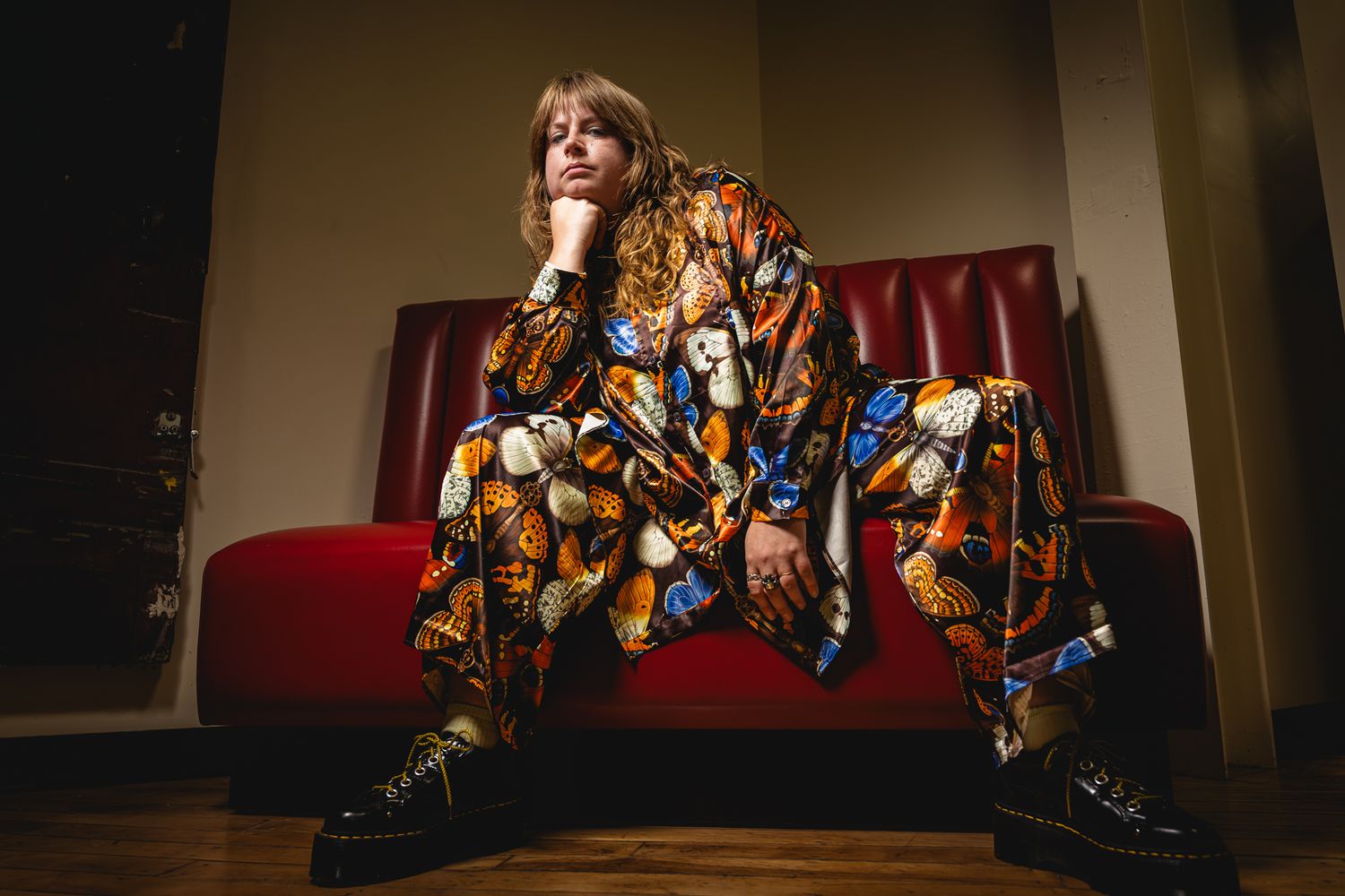 Person in colorful patterned jumpsuit and black boots sitting on red couch in moody lit room.