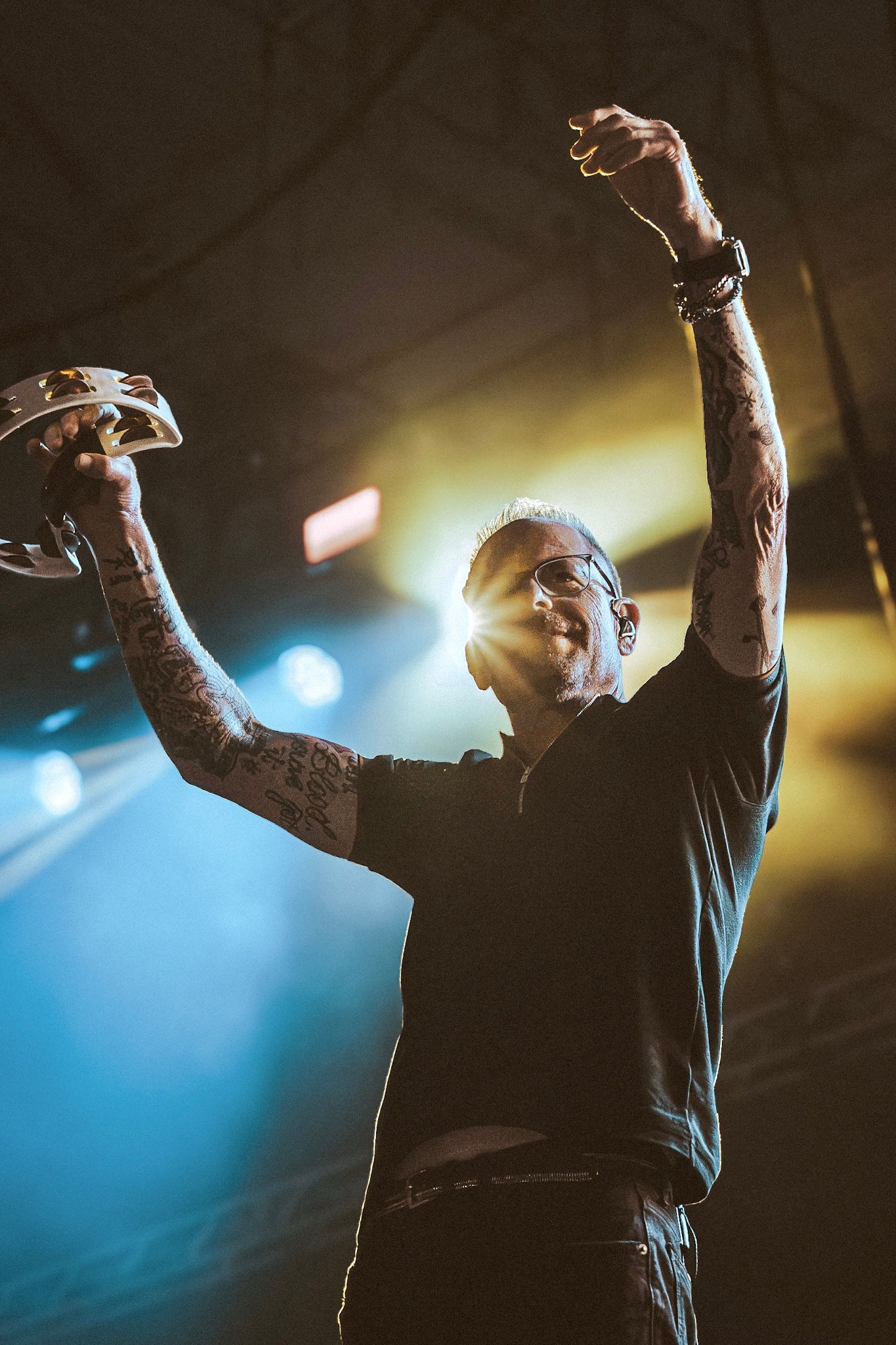 Tattooed performer raises arms triumphantly on stage, holding tambourine, illuminated by blue and yellow concert lights.