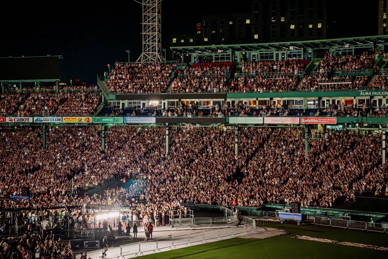 Massive crowd at a nighttime stadium concert with bright lights illuminating the stage and packed audience stands.