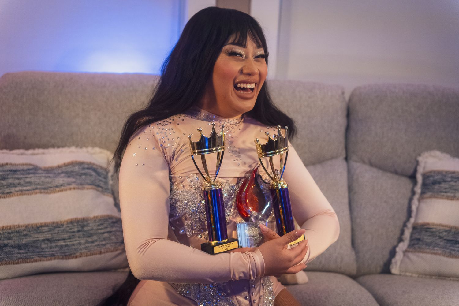 Someone wearing a white sweater sits on a grey couch holding two blue and gold trophy awards while laughing joyfully.