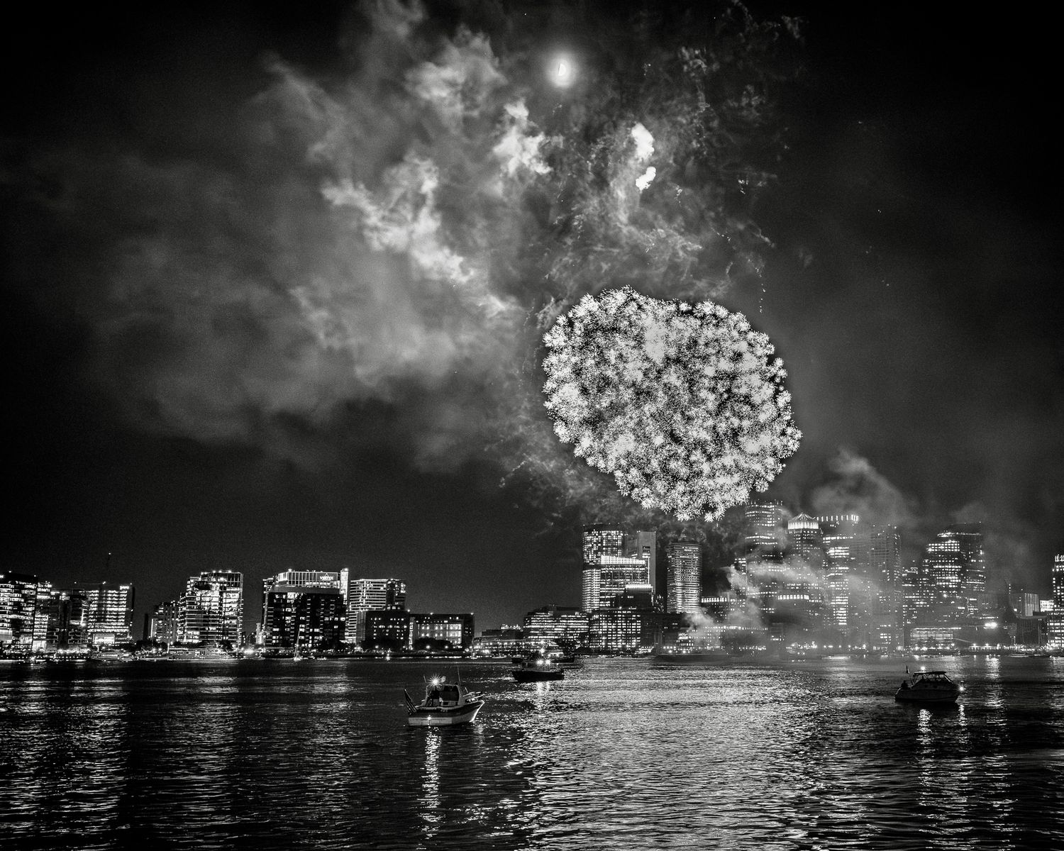 Black and white cityscape at night with fireworks bursting over illuminated buildings reflected in harbor waters.