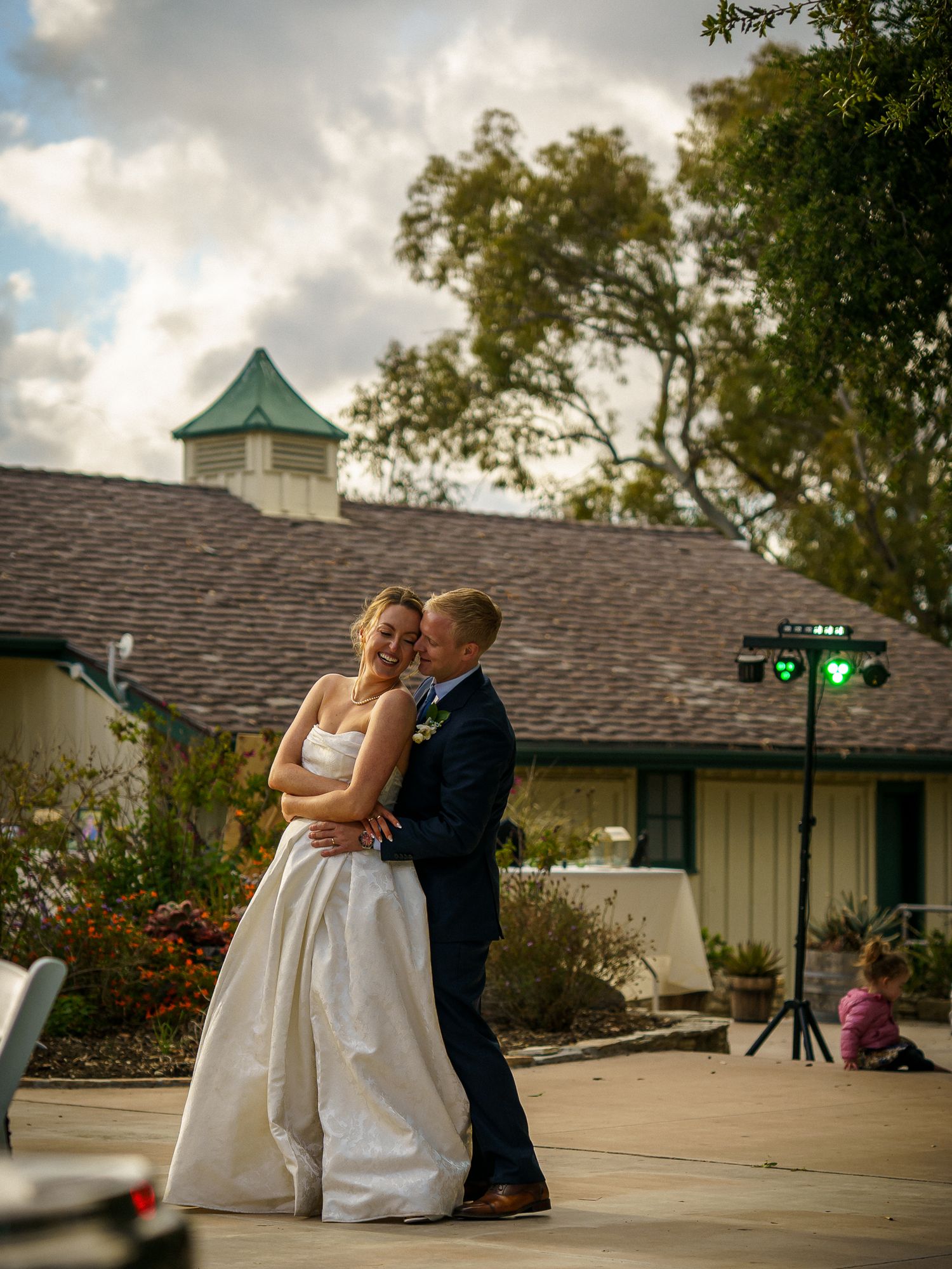 A couple shares a romantic moment on an outdoor dance floor at sunset with a white building and cupola in the background.