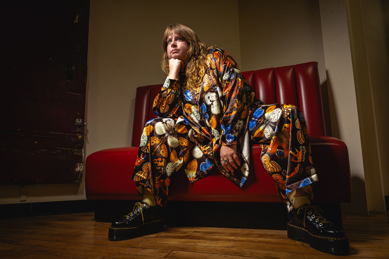 Person in colorful printed outfit sitting on red sofa with wooden floor background in artistic fashion photo.
