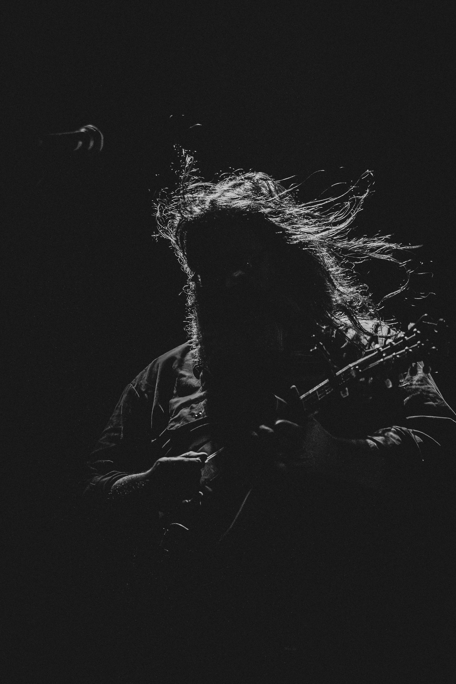 Black and white photo of a long-haired guitarist performing on stage with hair flying dramatically in dark lighting.