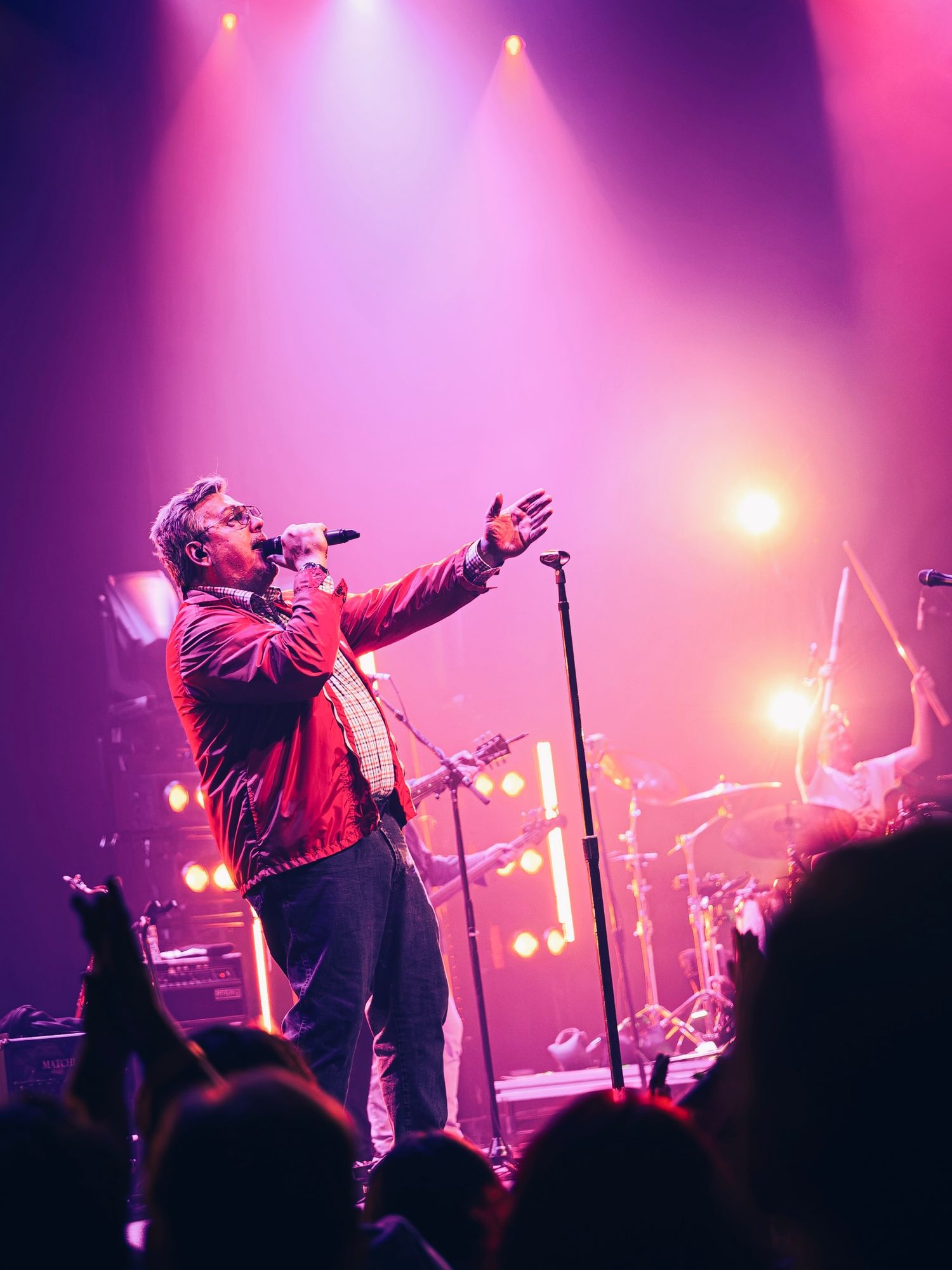 A male singer performs on a vibrant pink-lit stage, holding a microphone and gesturing to the crowd at a live concert.