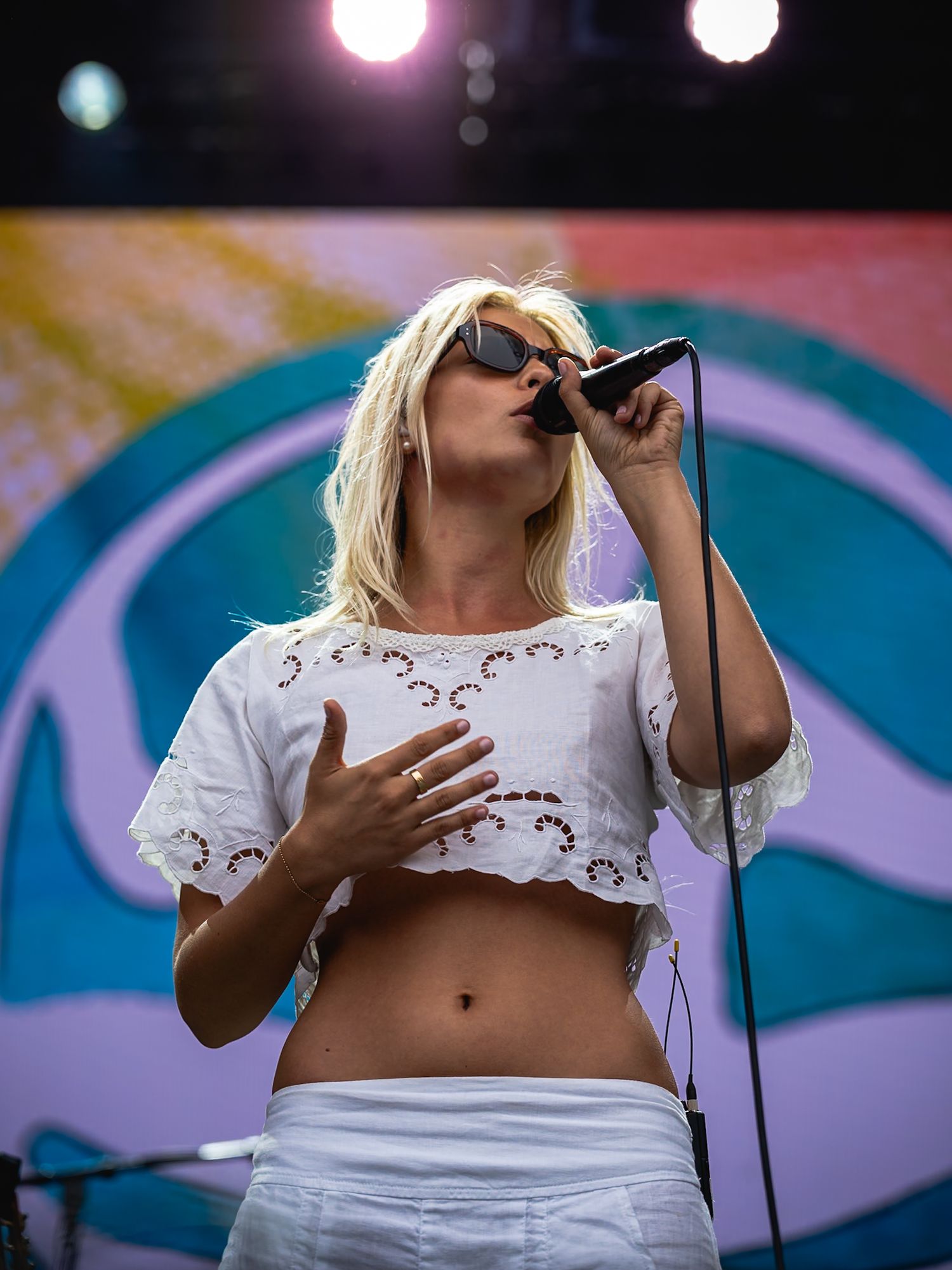 Young blonde female singer performing on stage in a white crop top, holding a microphone with colorful backdrop.