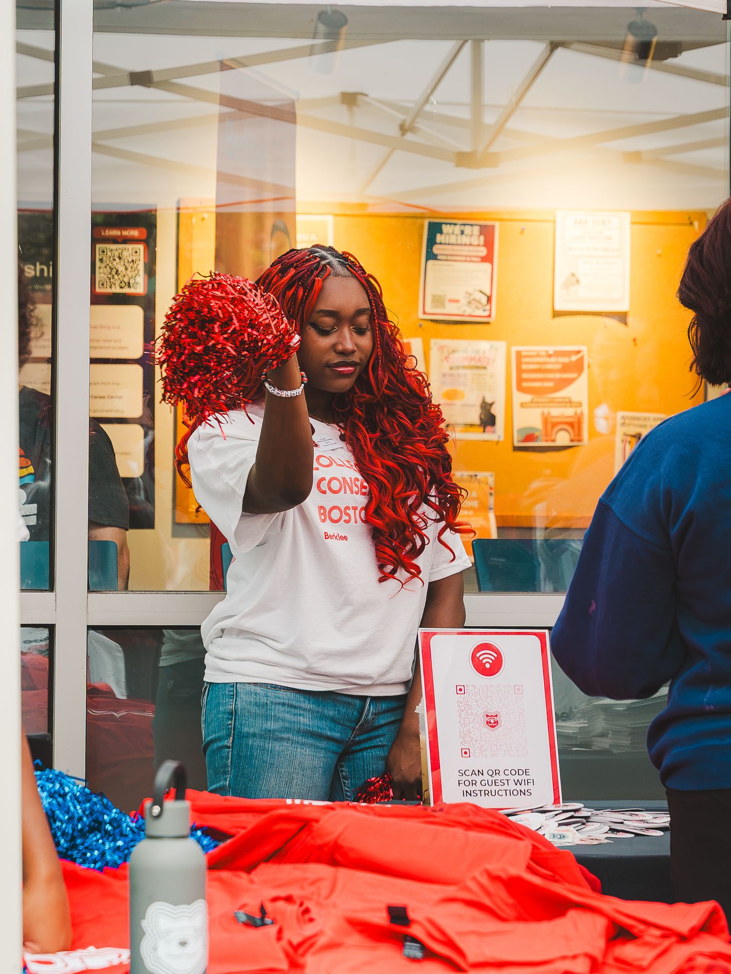 A young woman in a white Boston shirt holds red pom-poms at an event booth with red merchandise on the table.