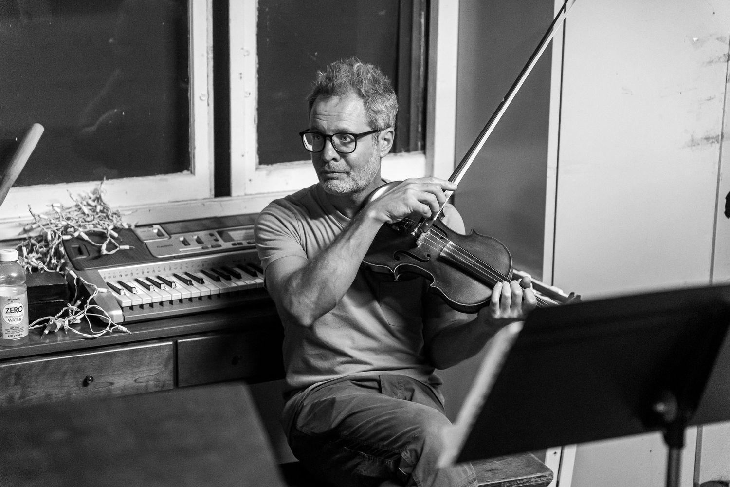 Black and white photo of a musician playing violin while seated at a music studio with keyboards visible in background.