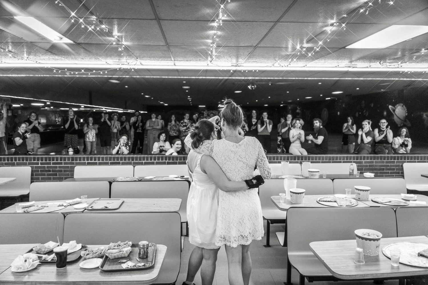 Black and white view of figures embracing at a roller rink table setup with a crowd in the background.