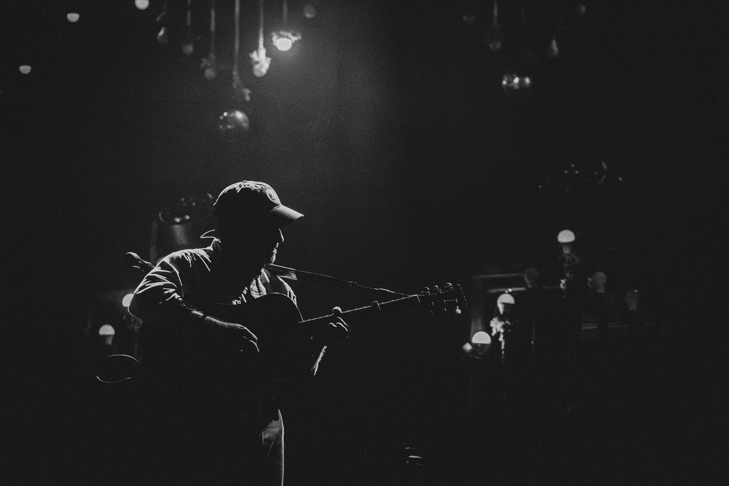 Black and white photo of a guitarist performing on a dimly lit stage under spotlights.